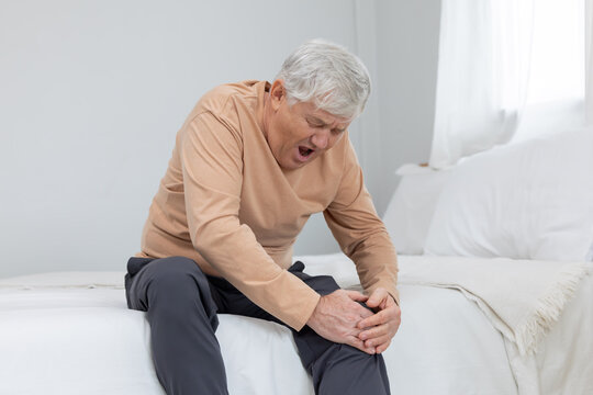 Older man sitting on bed holding his knee in pain during morning