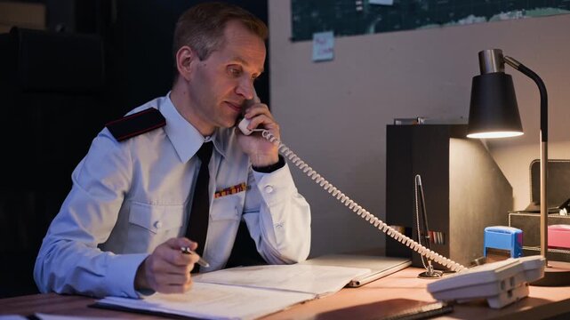 Middle aged Caucasian military officer talking on landline telephone and writing in logbook at desk in command center. Clip is suitable for government agency training or security service commercials