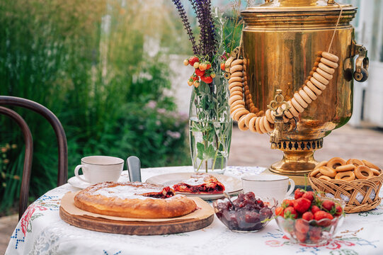 A rustic tea setting with golden samovar, fresh strawberries, jam, pie, and bagels arranged on an embroidered tablecloth outdoors.