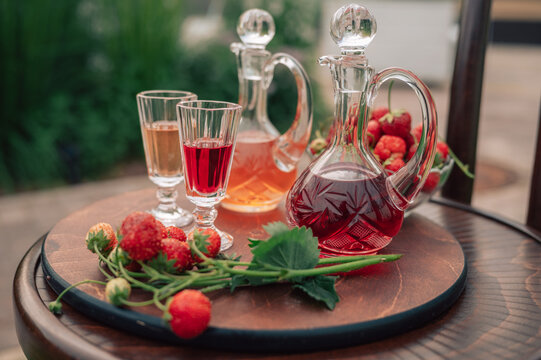 Crystal decanters with strawberry liqueur and glasses set on a wooden table, decorated with fresh strawberries in a natural garden setting.
