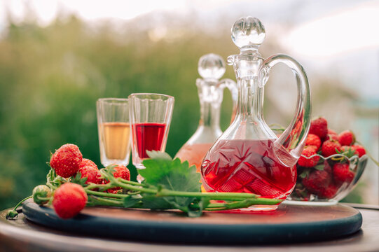 Crystal decanters with strawberry liqueur and glasses set on a wooden table, decorated with fresh strawberries in a natural garden setting.