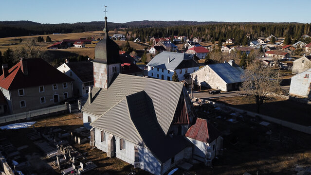 Aerial view of La Chapelle des Bois church surrounded by traditional village houses, a cemetery, and a forested landscape in Mouthe, Bourgogne-Franche-Comte, France.