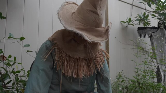 Burlap scarecrow with dripping black eyes stands by a white backyard fence. Spooky outdoor Halloween decoration in a garden during daylight.
