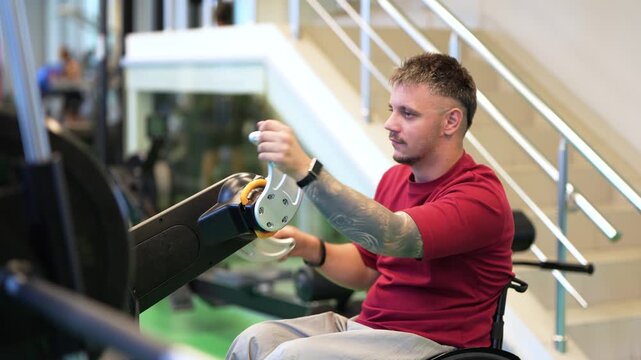 Motivated disabled man athlete in wheelchair at gym training using upper-body exercise machine, focused male sportsman with spinal injury doing rehabilitation workout on adaptive fitness equipment.