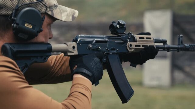 Shooter wearing hearing protection holds and aims a modern rifle during training at an outdoor shooting range.