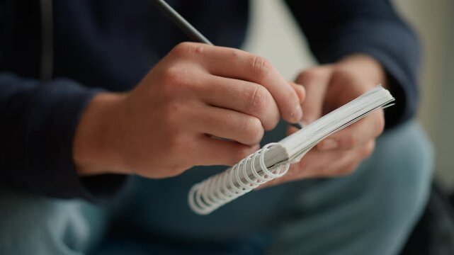 Close up of hands holding pen and spiral notebook during writing process. Notebook pages visible fingers gripping objects firmly. Soft indoor lighting with blurred background emphasizing