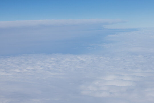 An aerial view from an airplane window looking out over a thick, rolling layer of low-level clouds beneath a clear blue sky. A vast sheet of white, textured clouds, typical of a low cloud layer.