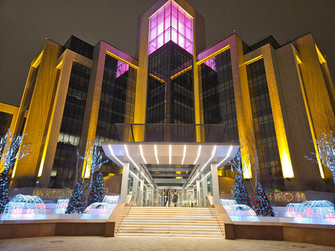 MOSCOW - DEC 08, 2025: Nighttime view of modern building LUKOIL with glowing purple and yellow lights. steps leading to entrance. Fluorescent trees and decorative fountains illuminate the plaza