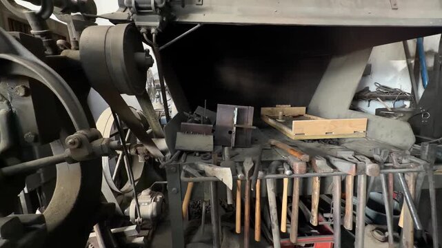 Interior of a traditional blacksmith workshop showing a belt driven power hammer, a forge, and a rack of various hammers and tools