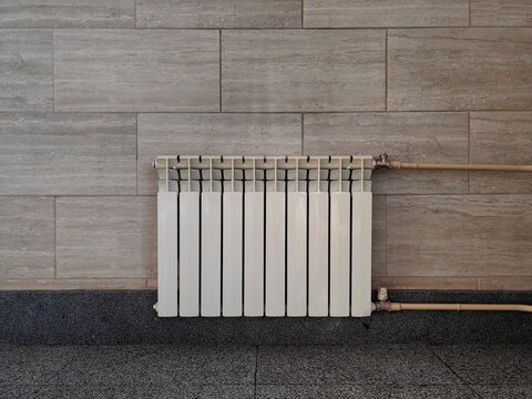 white metal section heating radiator pipes mounted on light beige travertine tile wall interior above dark grey polished granite floor surface, modern indoor temperature control appliance. vertical.