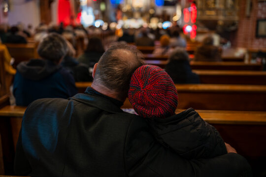 View from behind of a couple sitting closely in wooden church pews during a service, with the woman wearing a red knitted hat and leaning on his shoulder.