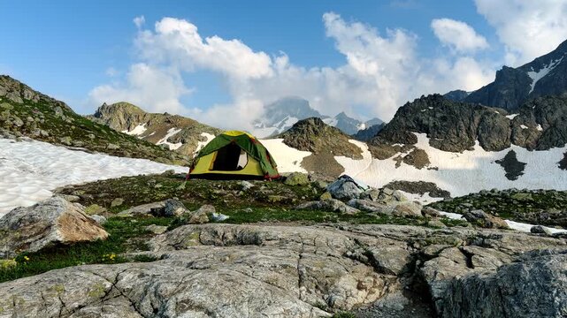 A yellow-green tent pitched on a rocky alpine ridge surrounded by snow patches, wildflowers and towering peaks, conveying high-altitude adventure, solitude and remote outdoor wilderness camping.