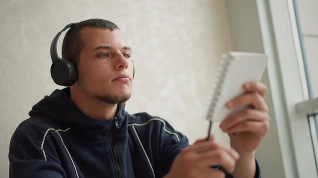 Close up of young man wearing headphones writing in notebook while holding pen near window with light colored wall background closeup adult indoor portrait