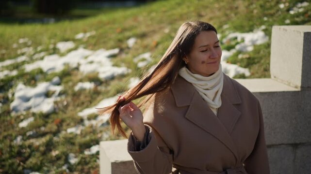Close up of woman adjusting her hair and neck in outdoor setting with grass patches of snow and concrete blocks. Woman wears beige coat over white scarf interacting naturally by touching
