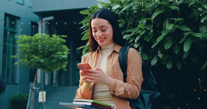 Beautiful young Caucasian woman with long hair standing outdoors, smiling while using a smartphone, with a modern glass office building in the background.