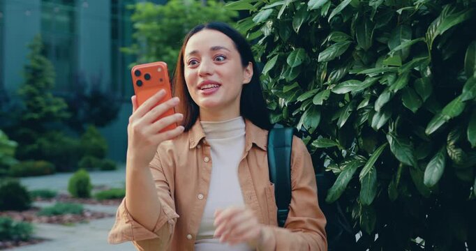 Joyful student woman on the street while holding a smartphone in front of her, engaged in an online video call with a friend. City buildings visible in the background.