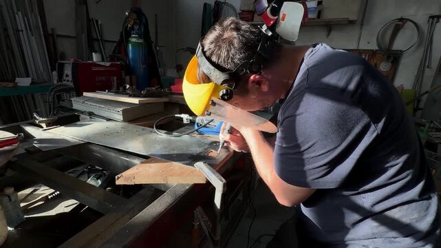 Focused craftsman wearing a protective face shield precisely engraving a metal plate. Intense light highlights the detailed work in the shop