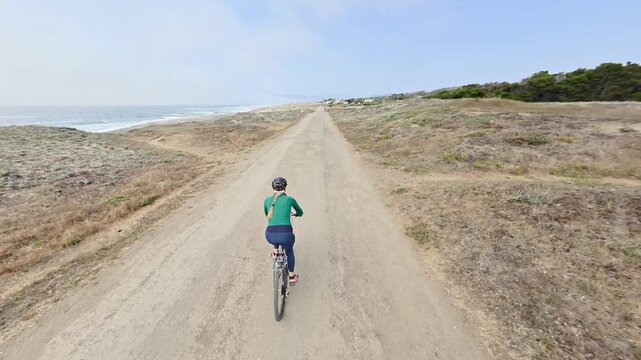 Mature woman rides her bike along paved trail in Fort Bragg, California by the ocean on a sunny day enjoying the outdoors.