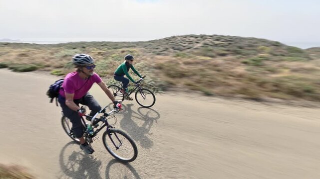 Mature man and woman bike along a paved trail and bridge in Fort Bragg, California with ocean views on a clear day.