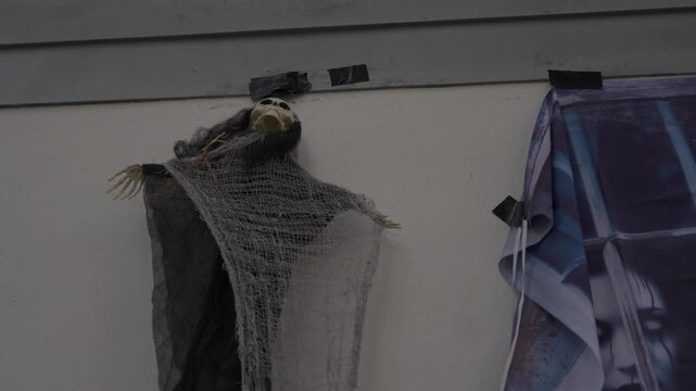 A hooded grim reaper with chains hangs by the house roof. A small skeleton ghost with gauze fabric is taped to the wall, creating a spooky Halloween yard decoration.