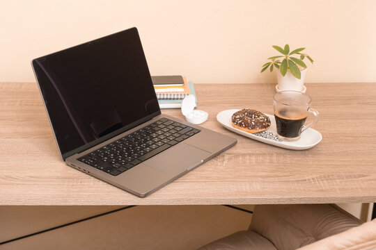 Laptop sits on a desk next to a donut and a cup of coffee in a home office setting during the morning