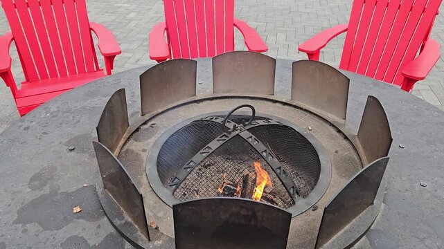 Red chairs surround a fire pit with active flames. Metal guard protects the fire from wind and debris. Fire burns steadily in a round, open pit. Surrounding surface shows signs of use and weather
