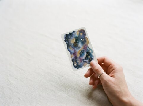 Woman hand holding tarot card with galaxy print. Cosmic watercolor nebula with stars, moon and planet. Fortune telling, astrology and esoteric divination concept.