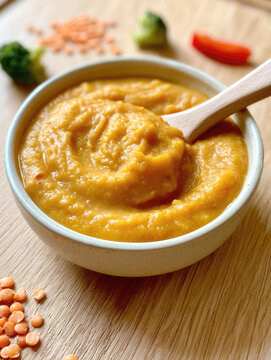Healthy organic pumpkin and red lentil puree soup served in a ceramic bowl with a wooden spoon, garnished with fresh broccoli, red bell pepper slices, raw lentils on a light wood tabletop background.