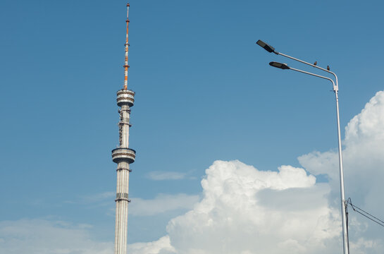 Low angle shot of a tall television broadcast tower standing near a street lantern against a clear blue sky with white clouds