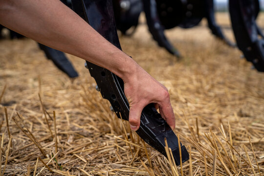 Farmer hand inspecting cast metal ripper tooth of agricultural subsoiler machine on dry wheat stubble field before deep tillage work.