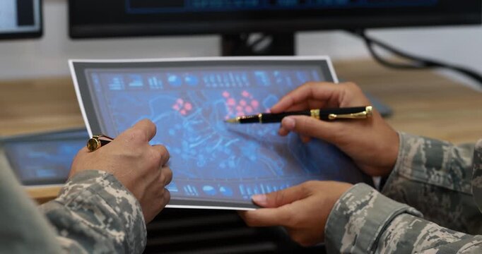 Two people in camouflage uniforms review a digital map on a tablet, using a stylus in a command center with computer monitors.
