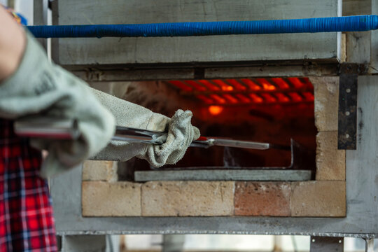 Artisan wearing heat resistant protective glove using metal tool to place workpiece into glowing hot muffle kiln during thermal firing in workshop.