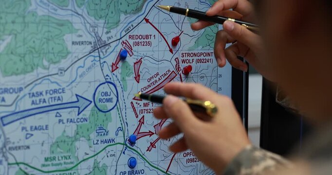 Military officer points at a tactical map with a pen, reviewing marked routes, targets and unit positions during mission planning.