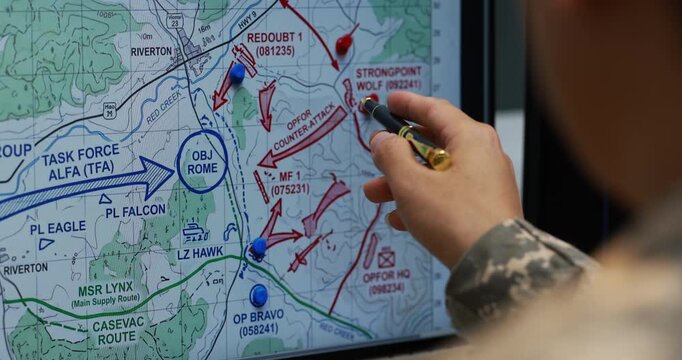Close-up of a person using a pen to point at a military planning map with marked routes, arrows, locations and tactical notes on a screen.