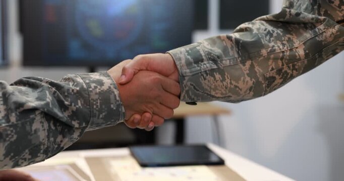 Close-up handshake between two people in camouflage uniforms across an office desk, with a map and tablet nearby, symbolizing military cooperation, trust, and agreement