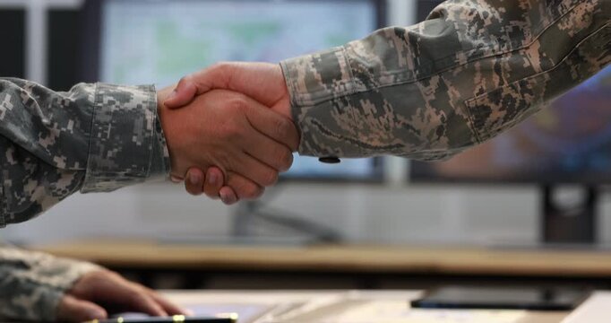 Military personnel shaking hands during a professional meeting, with computer monitors and planning materials in the background.