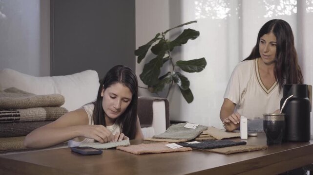Two women sit together at a wooden table, chatting and examining fabric and rug samples spread out before them in a bright