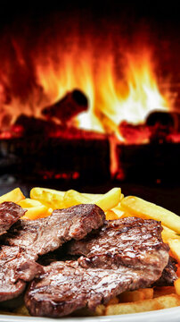 Juicy grilled picanha steak with golden french fries in front of a warm roaring fire. Traditional Brazilian barbecue food photography.
