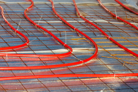 Red heating pipes lie beneath a floor grid. They are laid in a curved, overlapping pattern. The system is designed for radiant floor heating.