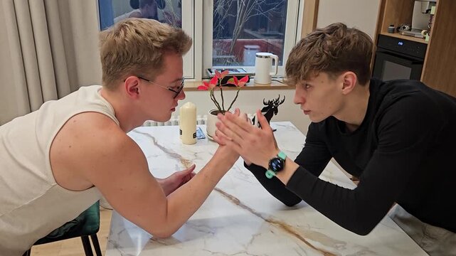 Two young men arm-wrestle at a marble table. One leans forward, muscles tensing. The other smiles, watching the match. Small black figurine sits nearby. Plants and a candle adorn the table