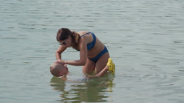 A caring mother gently helps her brave son float in the water. They laugh happily inside a calm lake filled with green algae.