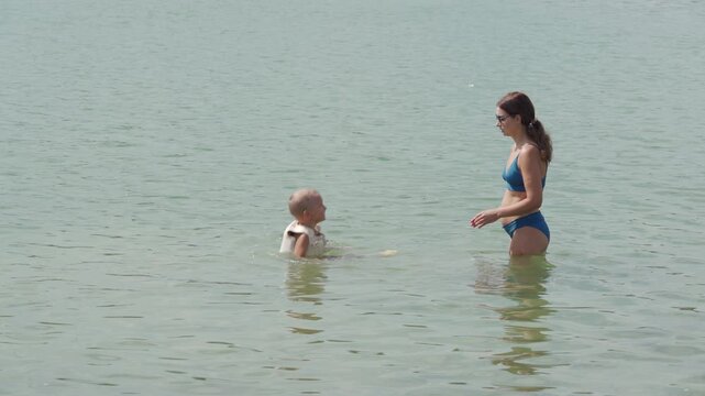 A happy boy and his loving mother swim together. They smile warmly inside a calm, greenish lake filled with microscopic green algae.
