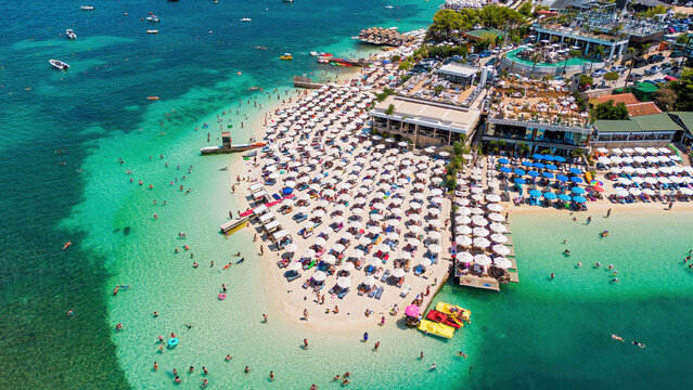 High-angle aerial drone view of the crowded beach, white umbrellas, and crystal-clear turquoise waters in Ksamil, Albania