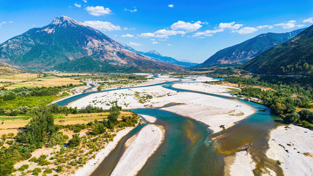 Aerial panoramic drone view of the wide braided riverbed, turquoise channels, and mountain peaks of the Vjosa River valley in Tepelen, Albania