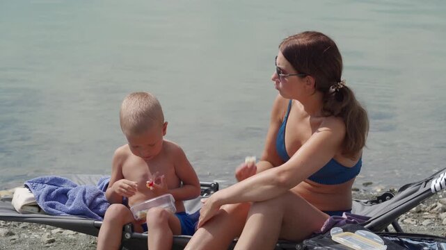 A loving mother and her happy son relax together on a beach cot. They share sweet snacks on a calm lakeside where green algae grows.