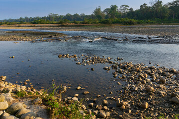 Naklejka premium Murti river in the morning, riverbed in foreground with flowing water and stones. Scenic beauty of Dooars landscape, West Bengal, India. Dooars is popular tourist destination for natural landscapes.