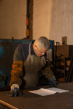 Hispanic industrial worker wearing protective clothing and gloves, with his hands resting on a workbench while carefully reviewing a technical blueprint in an industrial workshop
