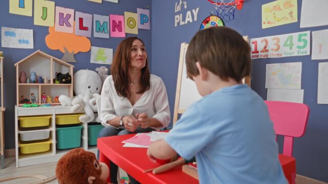 Woman leaning forward smiling helps little boy hold mallet and play xylophone in preschool classroom; joyful learning.