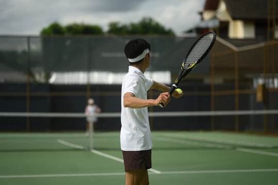 Rear view of Young Asian Man Preparing to Serve on Outdoor Tennis Court with Opponent in Background