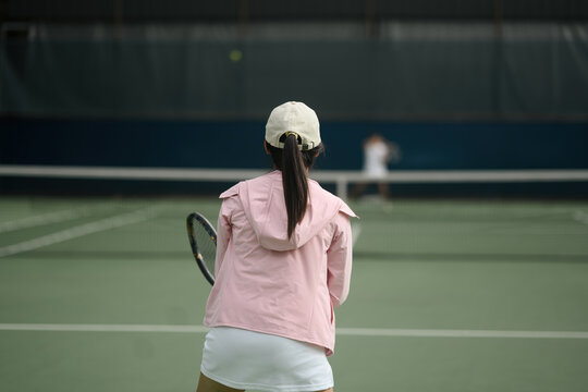 Young Asian Woman Playing Tennis on Indoor Court, Rear View with Racket and Pink Jacket